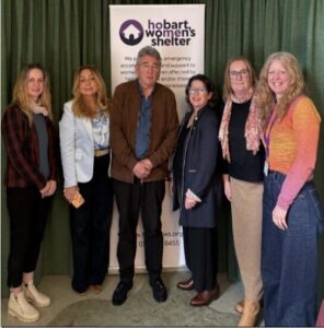 Six people posing together in front of a Hobart Women's Shelter banner at an outreach event, smiling for the photo.