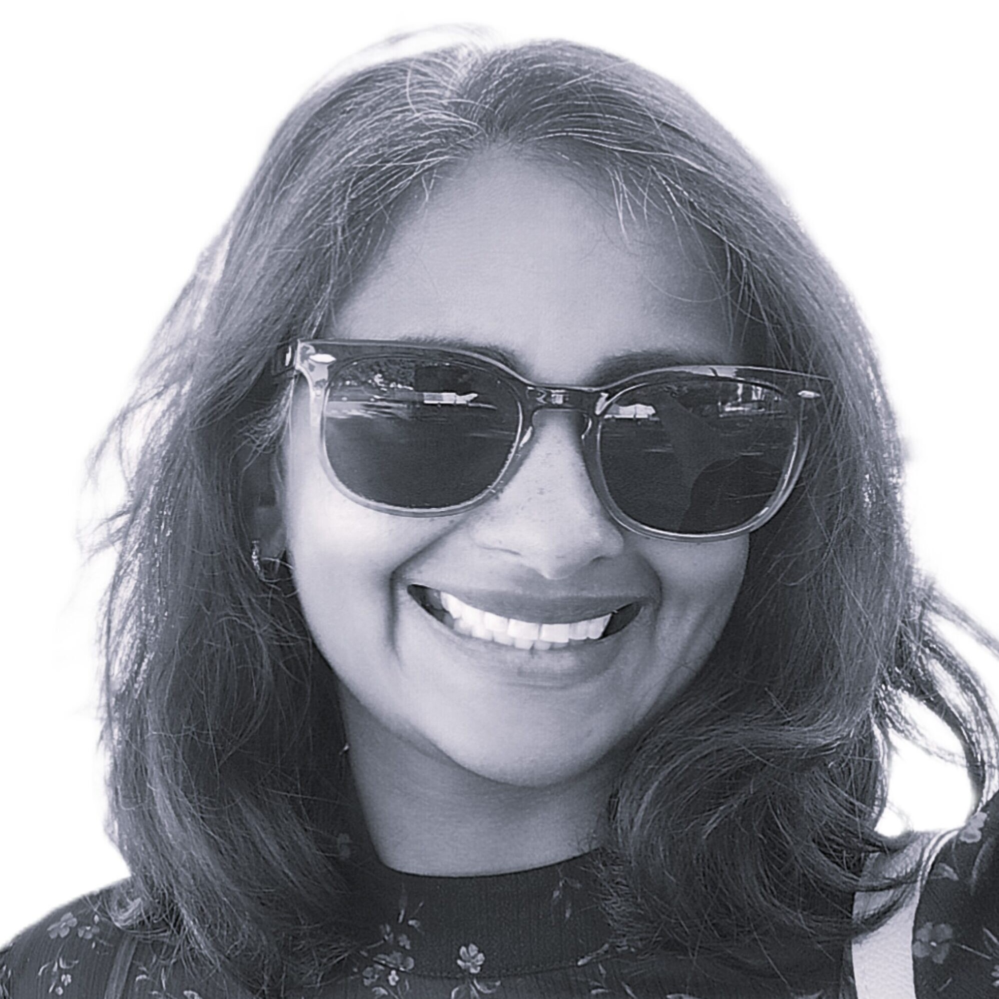 Smiling woman wearing dark sunglasses and a floral dress, with a light crossbody bag strap visible on her right shoulder.