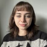 Portrait of a young woman with short brown hair and a nose piercing, wearing a black-and-gray sweater, looking at the camera.